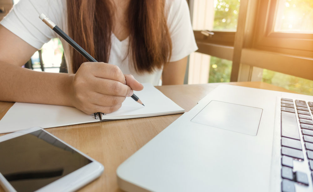Student writing design specifications by hand while researching with a laptop